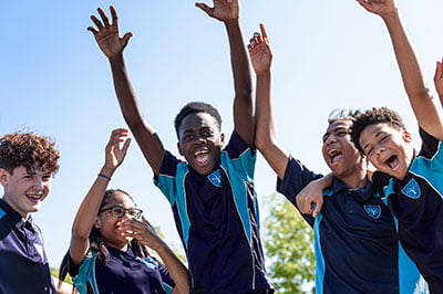 Students at King Edward VI Balaam Wood Academy smiling with raised hands outdoors.